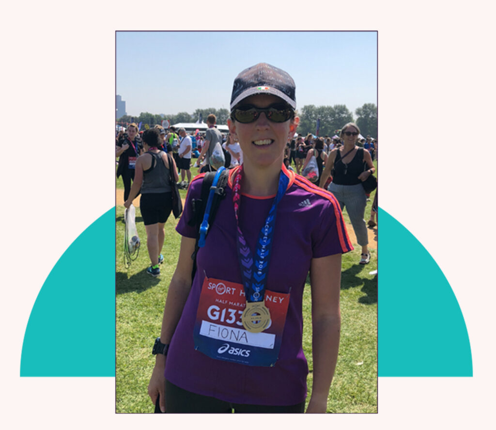 Photo of me after the Hackney Half Marathon wearing my race number and medal with a baseball hat, purple tshire and sunglasses. There are crowds in the background